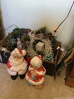 Santa and Mrs. Claus plastic figures standing on carpet with box containing wreath and decorations in background