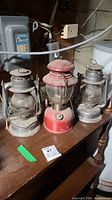 View of three lanterns including one red Coleman and two silver Beacon lanterns on a wooden table in basement setting.