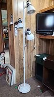 Full view of white metal floor lamp showing three adjustable light fixtures mounted on a tall vertical pole with round base. The lamp is positioned against a wood panel wall.