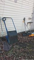 Full view of assorted garden tools including snow scoops, shovels and scraper against siding wall