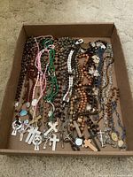 Image showing a flat box containing multiple rosaries of various colors and bead materials, cross pendants and religious medals visible.