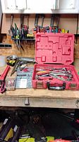Wide view of assortment of hand tools on workbench including red plastic case open with ratchets, wrenches and screwdrivers, various screwdrivers behind, and C clamps to side.