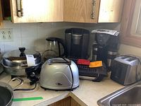 Wide view of two toasters, electric pot, electric glass kettle, two coffee makers, toast stamps, and electric grill on kitchen counter.