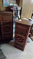 Pair of tall wooden speakers with decorative front grilles and visible wear on the cases, shown beside a bookcase and shelving unit.