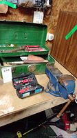 Overview of lot showing vintage green metal toolbox with hand tools inside, small black tool case on table, and blue bench vise mounted on workbench.