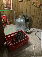 Wide shot showing the large glass jug, crate with empty beer bottles, and corker tools on the floor against a wood paneled wall.