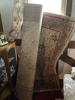 Photo of two rolled and partially standing area rugs shown indoors next to a cabinet and floral furnishings, rug with floral border design visible.