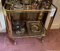 Full side view of the vintage metal framed bar cart showing glass shelves with silver and glass items on it.