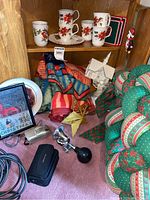 Photo of bottom shelves of corner cabinet showing Christmas mugs, plates, fabric angels, quilted wreath, throw blanket, framed Christmas print, bike horn, and Olympus camera.