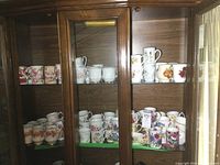 Two shelves inside a wooden cabinet containing various porcelain coffee mugs with floral patterns and a porcelain creamer.