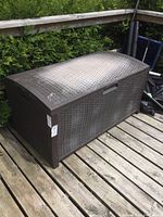 Full angled view of brown Suncast resin deck box on wooden deck, showing woven texture and some surface fading.