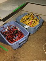 Two blue plastic totes on floor, each filled with multiple coiled extension cords, some heavy duty, variety of colors.