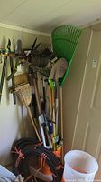 Full view of garden tools hanging and leaning including rakes, shovels, pitchfork, broom, hand trimmers, and various handles visible