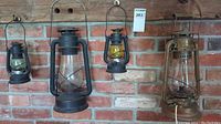 Three oil lanterns hanging against a brick wall, two smaller and one larger with wire handles.