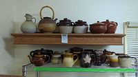Wide shot showing upper shelf with ceramic pitcher and bamboo-handled teapot, lower shelf with mugs and Brown Betty pots.
