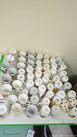 View of about 50+ mugs arranged on a table, showing tops and handles, mostly white with floral decorations.