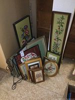 Photo showing multiple framed needlepoint pieces in various frame styles and sizes leaning against each other on carpeted floor with a metal display stand behind them.