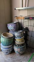 Seven ceramic planters of various sizes and designs stacked next to metal lattice and decorative stakes, with a green plastic edging strip on floor.