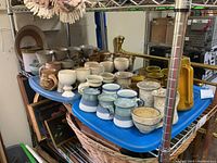 Wide view showing multiple rows of assorted pottery mugs, bowls, and pitchers on a blue tray with metal shelving surrounding them