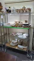 Full view of metal wire shelving unit loaded with various dishware and ceramic items. Four wire shelves, each holding multiple plates and pottery items, metal posts, and wheels visible.