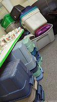 Stack of assorted plastic storage totes in various colors - gray, green, purple, clear, blue, beige, some with lids or handles, visible signs of use and dirt.