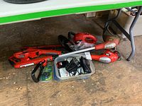 Photo shows two Black and Decker cordless weed trimmers and one hedge trimmer with multiple batteries and two chargers arranged on the floor beneath a table.
