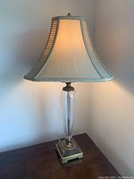 Side table lamp made of bronze and vertically ribbed clear glass with beige fabric shade, lit and photographed on a wooden surface.