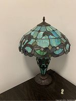 Antique style stained glass lamp positioned on dark wood surface, showing the front and side of the lamp, highlighting its colorful stained glass pattern and metal base.