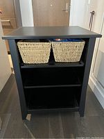 Front view of the black wooden shelf showing two wicker baskets on the top shelf and two open shelves below.
