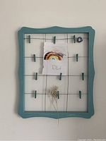Photo showing the blue wooden picture frame with multiple green clothespin clips attached to wire grid for holding photos or papers.
