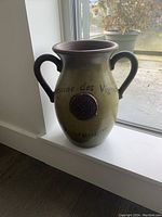 Green ceramic vase with two handles and embossed emblem placed on a white window sill with natural light.