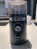 Front view of the Hamilton Beach coffee grinder showing the clear plastic lid, brand name, adjustable grind dial with ON/OFF button, and cup capacity slider.