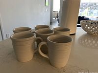 Set of six white coffee mugs arranged on a countertop with slight ribbed texture and large handles.