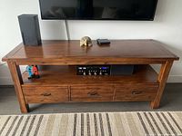 Wooden entertainment cabinet with a warm brown finish, holding electronic devices on the open shelf and three drawers below.