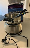 Electric fondue pot assembled and plugged in, viewed from side, showing stainless steel pot with two black handles and fondue forks resting inside with colored tips.