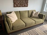 Front view of olive green three-seat sofa with two beige throw pillows, showing wood base