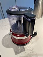 Front side view of the KitchenAid food processor showing the red base, black handle, and clear plastic bowl and lid.