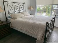 Photo showing a queen size metal bed frame with wrought iron and copper finish, set up with mattress and bedding, near a window and side tables with lamps.