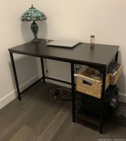 Photo of the dark wood office desk with black metal frame placed in a corner against two walls. Side shelving with two woven baskets visible on the right side under the desk surface. Desk has a decorative Tiffany-style table lamp, laptop, and hourglass on top.