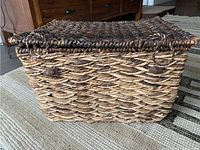 Photo of a small rectangular wicker basket with lid, showing the textured, interlaced natural brown wicker weave and wooden bead accents on the front.