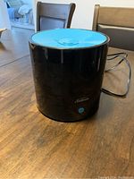 Black cylindrical Sunbeam humidifier with a blue top and power cord, placed on a wooden surface.