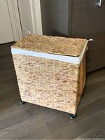 Wicker laundry basket in closed position showing woven texture, lid, and black wheels on floor.