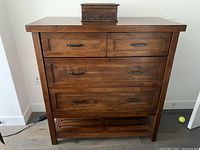 Front view of wooden dresser showing four drawers with metal handles, maple stain finish, and slatted bottom shelf. Small hole on back panel not visible.