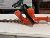 Side view of orange hedge trimmer with black handle and guard, showing 16-inch dual cutting blades.