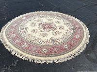 Round wool area rug 8-foot diameter with fringed edges, featuring cream, beige, red floral design, lying on black asphalt surface.