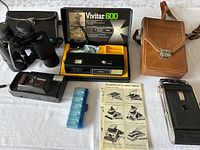 Overview of all items: Vivitar 600 camera in original box, Safari Starshot 35mm camera, Polaroid SX-70 Land camera with manual, black binoculars with case, and blue battery holder.