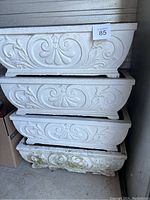 Four white decorative concrete planters stacked, showing raised scroll motif and soil