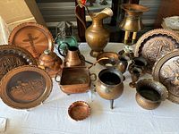 Photo showing various copper decor items including plates with embossed designs, pitchers, bowls, and other containers on a white tablecloth.