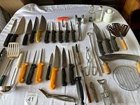 Full assortment of knives, kitchen tools, and utensils laid out on a white tablecloth.