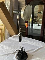Photo of an adjustable black metal desk lamp on a table with a white tablecloth, showing the round base, adjustable arm, and round metallic lampshade.